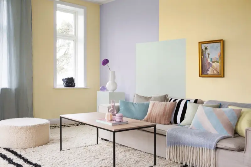 Ett vardagsrum med pastell inredning. Ljusgula, ljuslila och ljusgröna väggar skapar en mjuk atmosfär. En beigefärgad soffa är fylld med kuddar i olika pastellfärger och mönster.